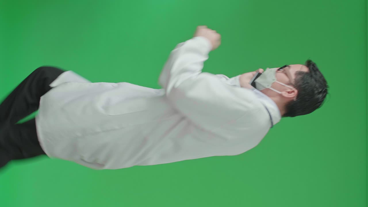 The Side View Of Asian Doctor With Stethoscope Wearing Mask, Glasses, And White Coat Talking On Mobile Phone While Running On Green Screen Chroma Key.