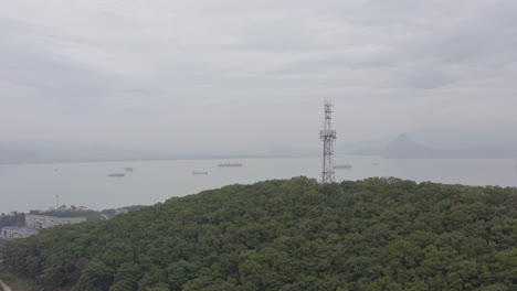 radio telecom tower on top of a green forest mountain with the reveal of bay area with ships at anchorage and mountain ridge in far distance