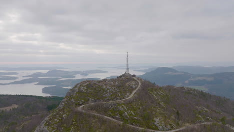 Camino-Sinuoso-Hasta-Una-Montaña-Con-Torre-De-Mástil-De-Teléfono-Con-Vistas-A-Un-Lago-Brumoso