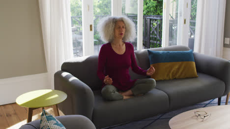 senior caucasian woman practicing yoga while sitting on the couch at home