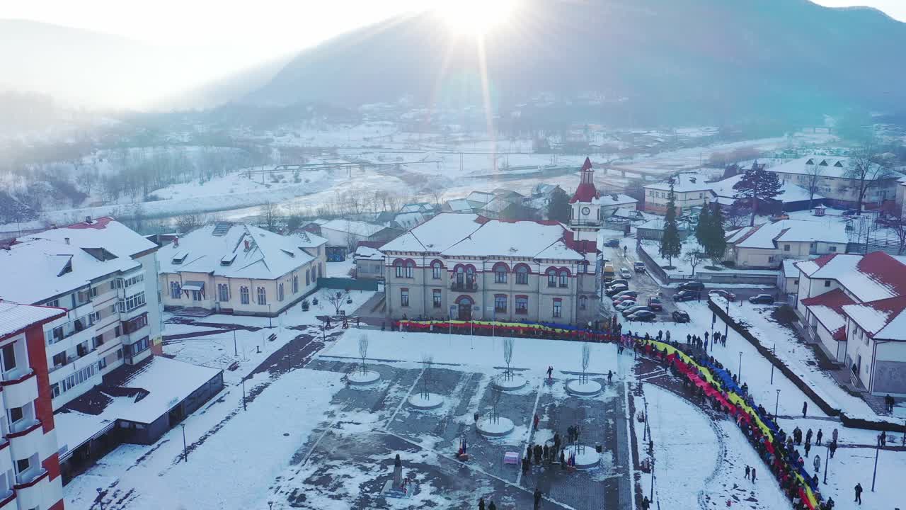 High-angle aerial shot of the snow-covered town square in Targu Ocna, Romania. A very long Romanian national flag is held by a line of people next to the historic town hall on a bright winter day