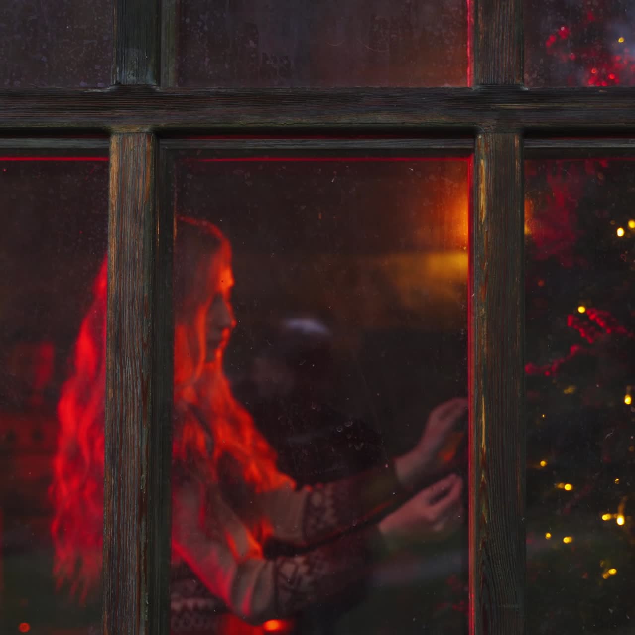 Comfortable atmosphere inside the house. Young family prepares for New Year. Woman decorates Christmas tree near the window. Husband brings hot tea. View from outdoors.