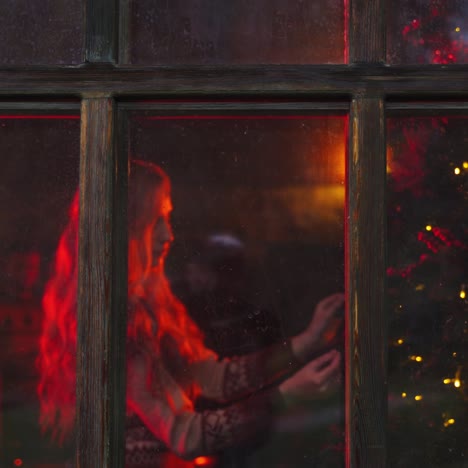 Comfortable atmosphere inside the house. Young family prepares for New Year. Woman decorates Christmas tree near the window. Husband brings hot tea. View from outdoors.