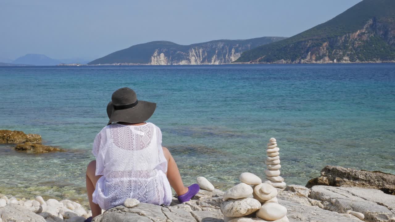 Female Tourist Sitting At The Rocky Shore Of Emplisi Beach In Erisos, Greece. wide shot