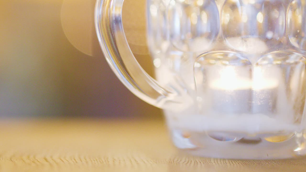 A softly glowing candle burns inside a clear glass mug on a wooden table, with a blurred, ambient background and warm lighting