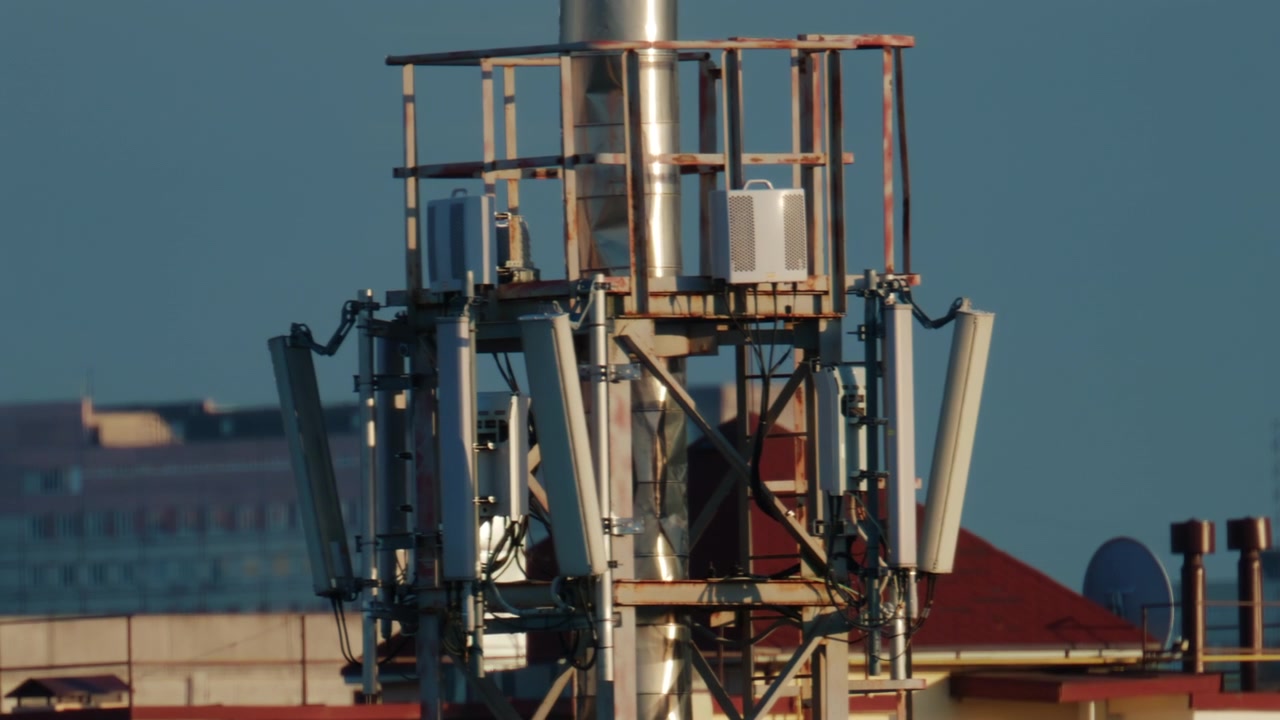 Scena su un tetto urbano che mostra una torre di comunicazione cellulare con antenne a pannello multiple, montata su una struttura metallica sopra edifici residenziali