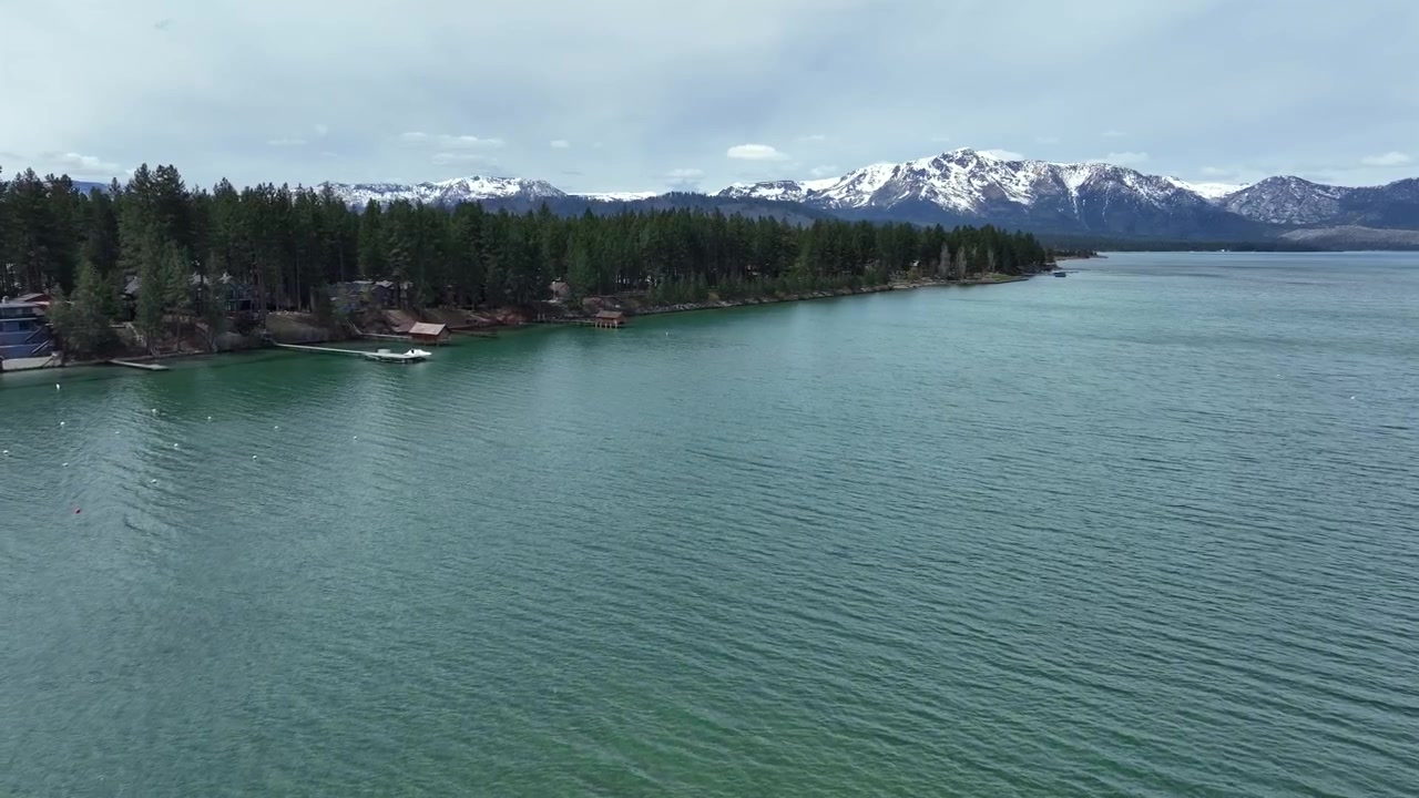 Acque limpide e turchesi del Lago Tahoe meridionale con foreste sempreverdi e montagne innevate della Sierra Nevada sotto un cielo nuvoloso in California, Stati Uniti. Ripresa aerea con drone.