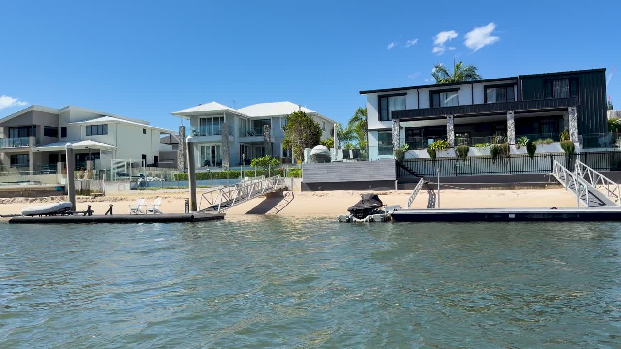 Camera glides past modern riverside houses, docks, and lush greenery under bright midday sunlight