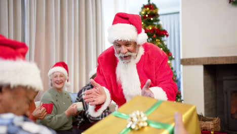 felices y diversos amigos mayores con trajes de papá noel y sombreros de navidad intercambiando regalos en casa, en cámara lenta