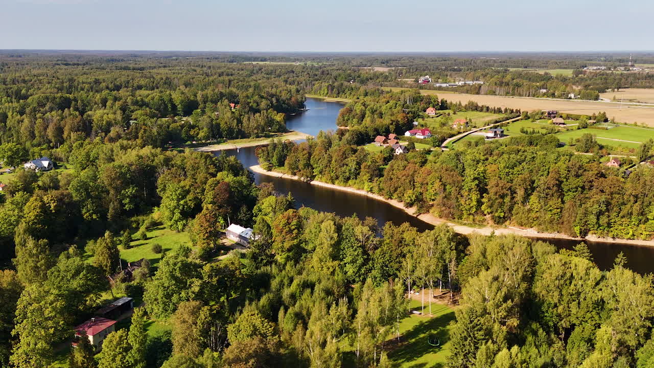 Aerial pullback zoom out from of buildings by river with lush greenery and winding shoreline in Latvia