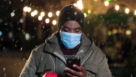 Portrait of african american man typing on smartphone and holding a christmas tree on the street while it¬¥s snowing in Christmas