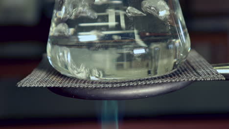 primer plano de la parte inferior del agua hirviendo en un matraz erlenmeyer en un soporte de anillo calentado por mechero bunsen