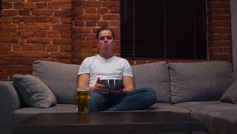 El-Joven-Está-Sentado-En-El-Sofá,-Viendo-La-Televisión-Y-Comiendo-Papas-Fritas-En-Su-Sala-De-Estar