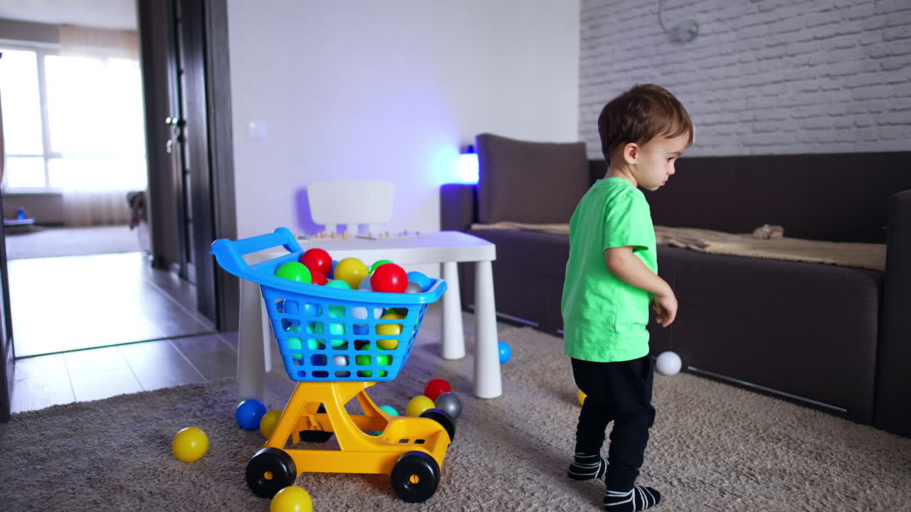 Sweet toddler boy touches colorful balls in the toy shopping cart. Cheerful baby turning around happily.