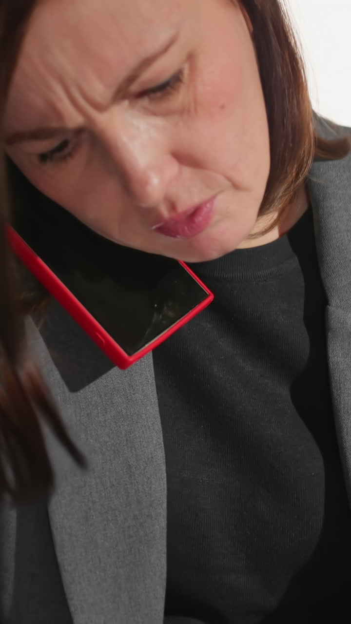 Businesswoman in gray suit holds phone between shoulder and ear while reading document with serious expression, multitasking in modern office setting, reflecting work stress, urgency