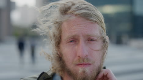 close up portrait young blonde hipster man student looking serious running hand through hair wind blowing in city background