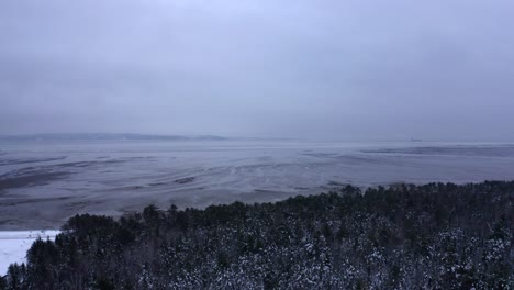 Drone-flying-over-a-snowy-forest-beside-the-Saint-Lawrence-river-in-Charlevoix