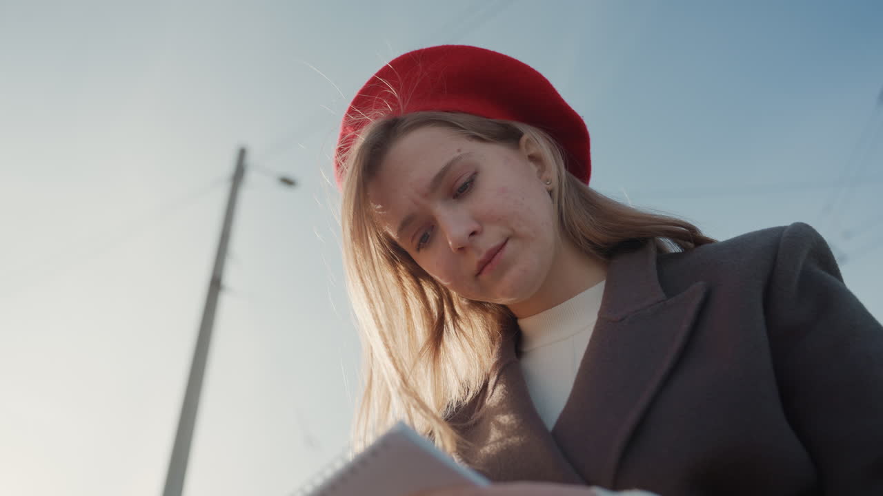 Giovane donna con berretto rosso che scrive un diario su un argine erboso accanto a un sentiero, penna sulla carta, espressione pensierosa, cappotto elegante, sfondo con palo del telefono, atmosfera cinematografica diurna per contenuti di studio.