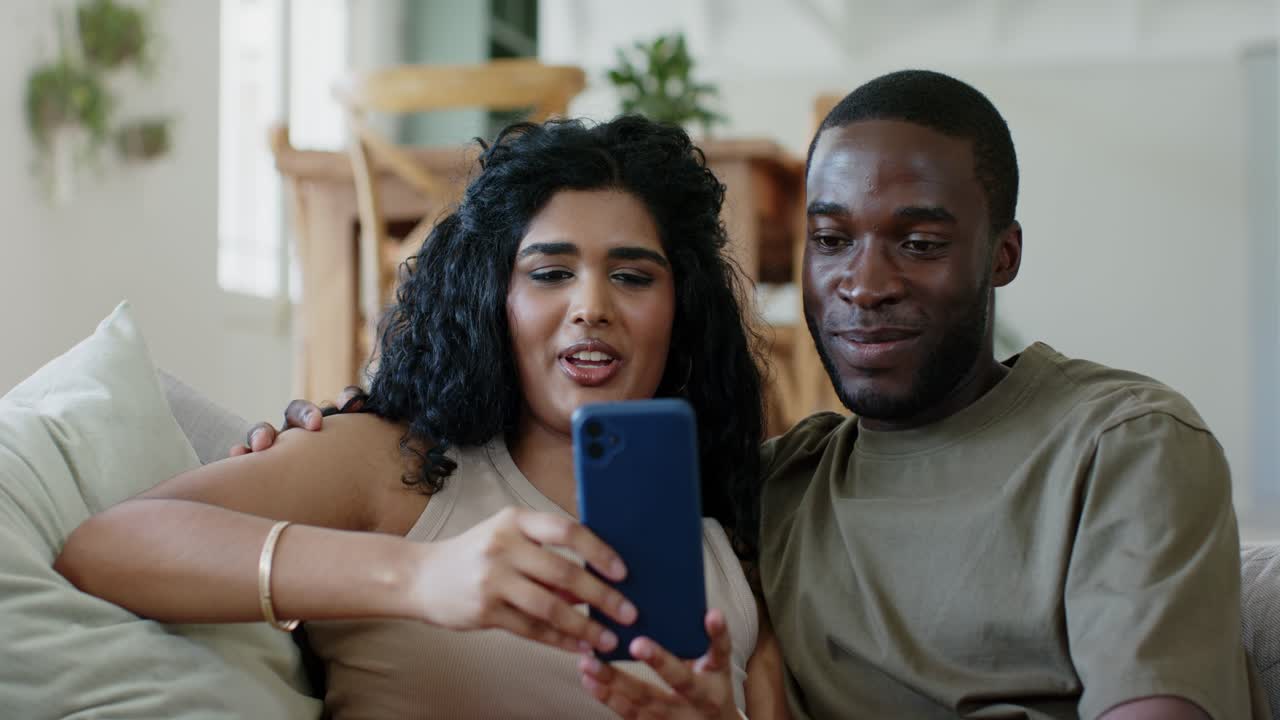 Diverse couple sitting on sofa in living room, woman tapping blue smartphone showing content