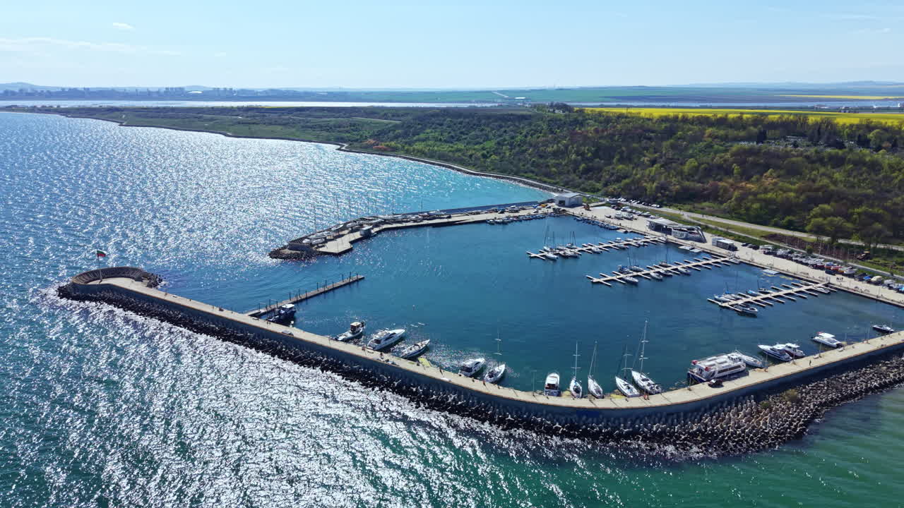 Udsigt over en marina med både og yachter på en solrig dag ved vandfronten i et kystområde nær et grønt landskab og blå himmel.