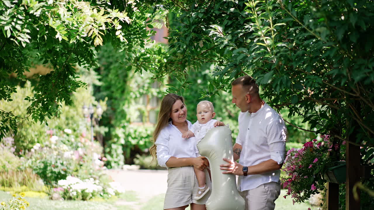 Beautiful young Caucasian family with baby boy. Happy parents give the kid a balloon in number one shape. Green garden at backdrop.