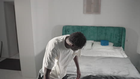 man in white t-shirt walking through minimalist bedroom carrying a backpack, looking down, with neatly made bed and green headboard in the background, preparing to leave, calm and simple environment