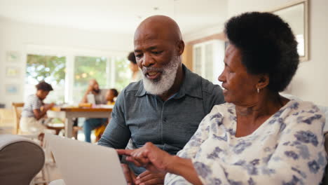 Senior-Couple-At-Home-Looking-At-Laptop-Together-With-Multi-Generation-Family-In-Background