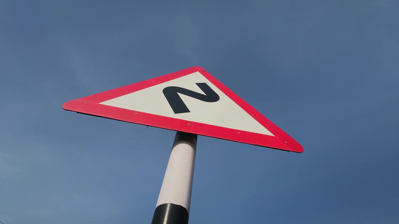 Camera orbiting around a triangular road warning sign indicating a sharp curve ahead, captured against a clear blue sky, highlighting transportation safety, navigation, and roadside infrastructure.