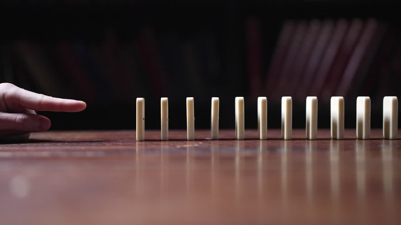 A finger pushes a line of white dominos, triggering a chain reaction in slow motion and showcasing the domino effect