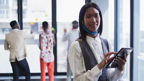 portrait of asian woman with face mask around her neck using digital tablet at modern office