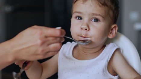 mom shoves a full spoon to her son but he turns away. kid takes the food and holds it in his mouth. close up portrait.