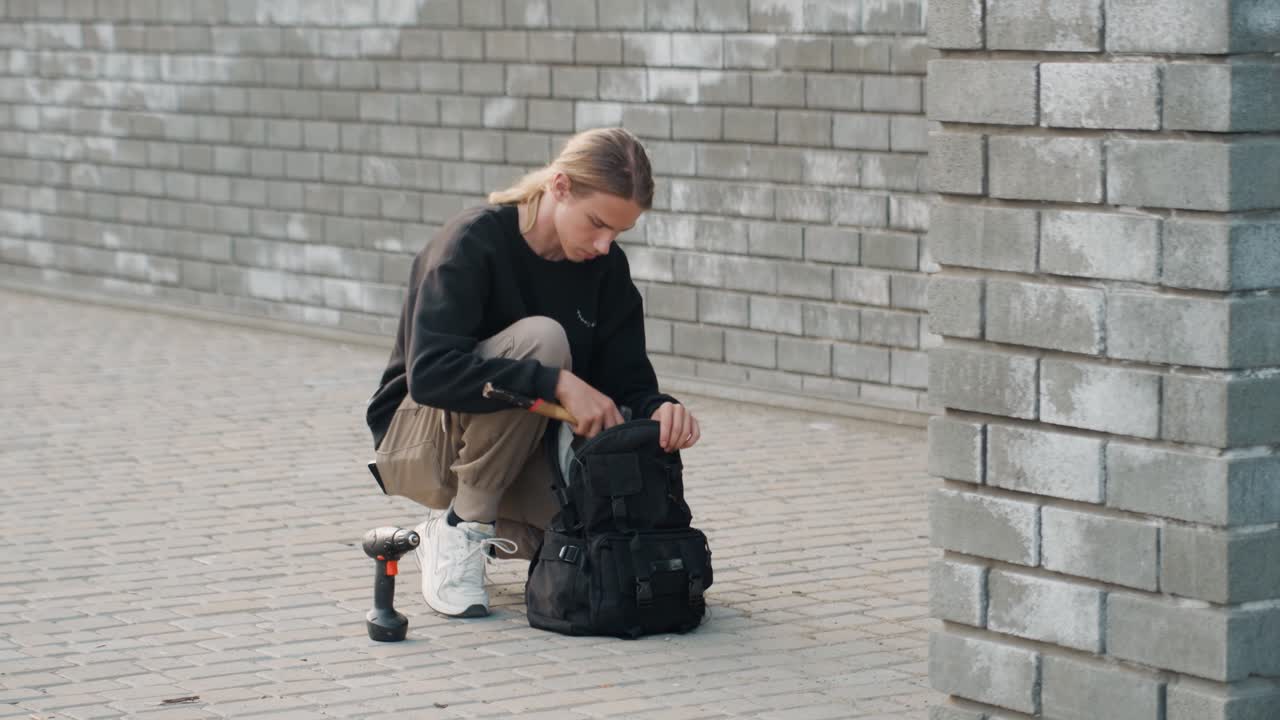 Młoda osoba klęcząca przy plecaku, sprawdzająca gimbala i sprzęt fotograficzny na tle ceglanego muru, ciche miejskie otoczenie, skupione przygotowanie do sesji ulicznej, subtelne światło i teksturowany chodnik.