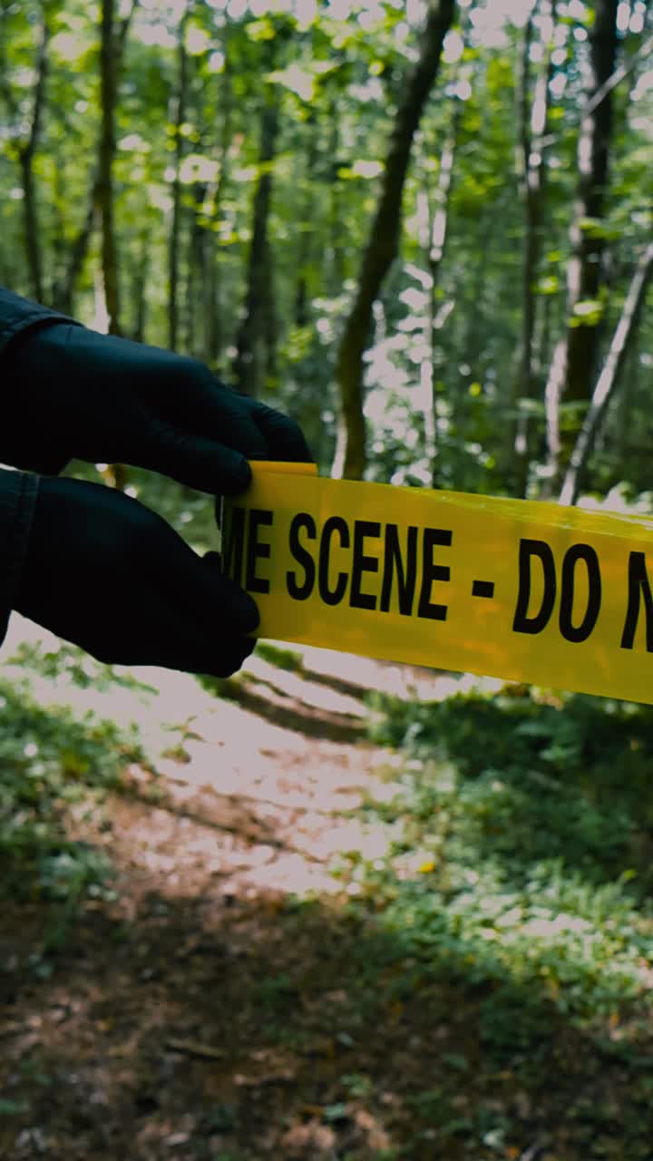 Close up vertical video of a yellow colored Crime Scene Do Not Enter police caution tape ribbon being pulled in front or unraveled by investigator hands that have black gloves on in a sunny forest