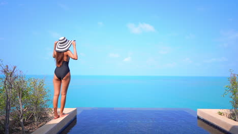 Pequeña-Mujer-En-Traje-De-Baño-En-Tonos-De-Piscina-Infinita-Azul-Y-Agua-De-Mar-Tropical-Bajo-Un-Cielo-Despejado-De-Verano-Levantando-Las-Manos-En-El-Aire,-Cámara-Lenta