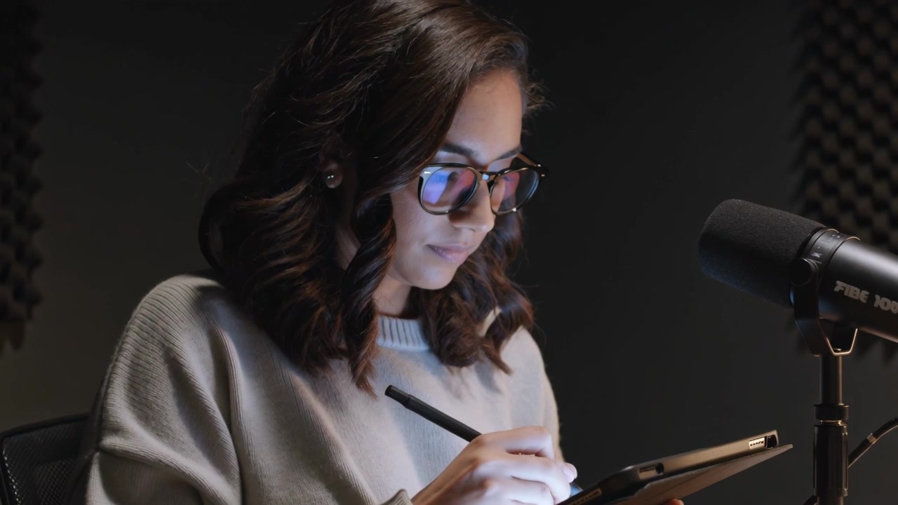 Donna con maglione e occhiali che disegna tenendo uno stilo su un tablet, dando un'occhiata al microfono in una cabina di registrazione.