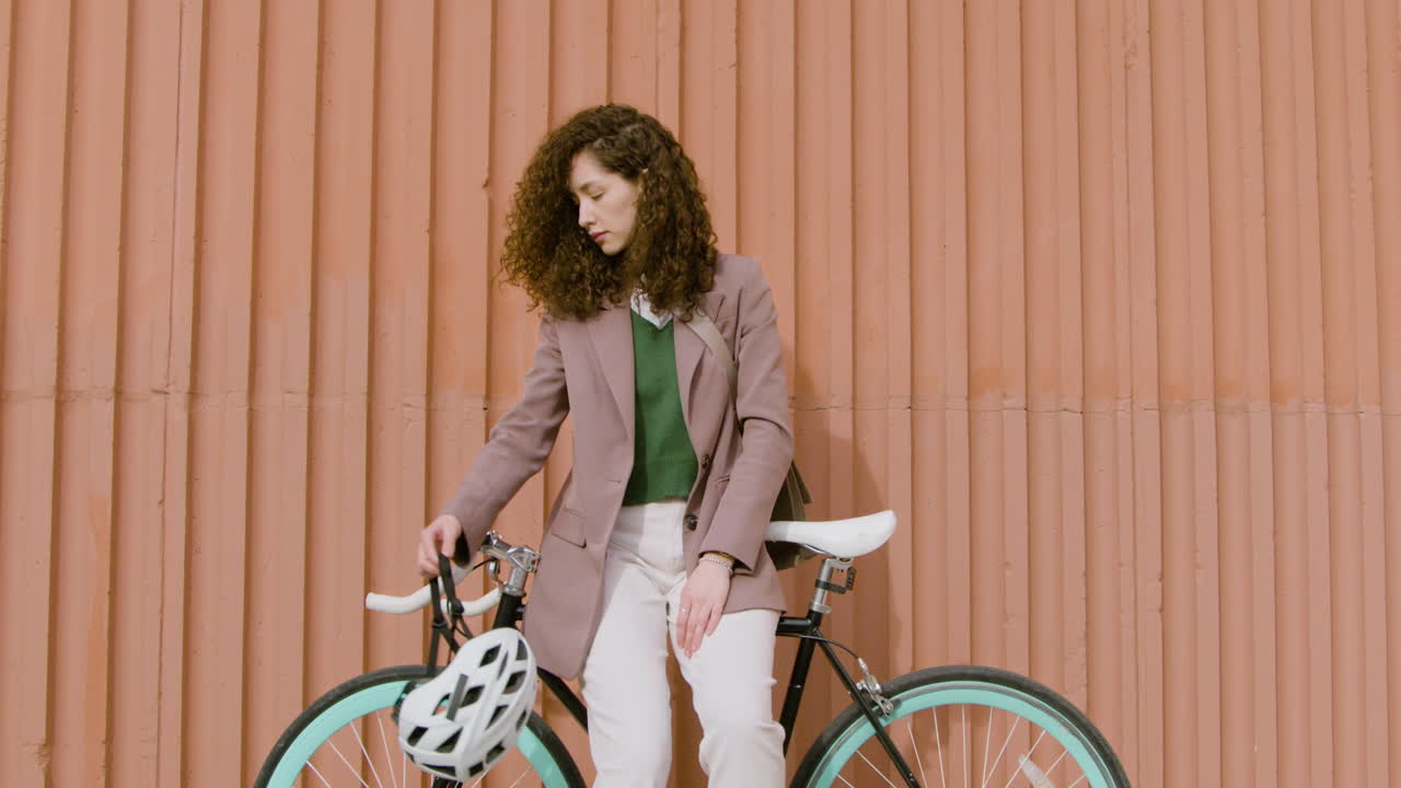 niña rizada sonriente con ropa formal mirando la cámara mientras se apoya en bicicleta frente a un edificio de metal prefabricado