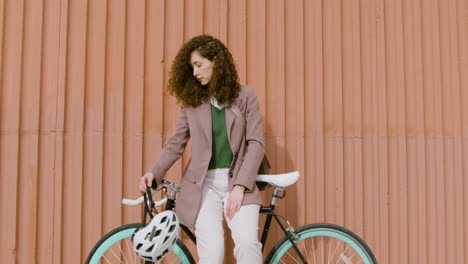 menina encaracolada sorridente em roupas formais, olhando para a câmera enquanto se apoiava na bicicleta em frente a um prédio de metal pré-fabricado