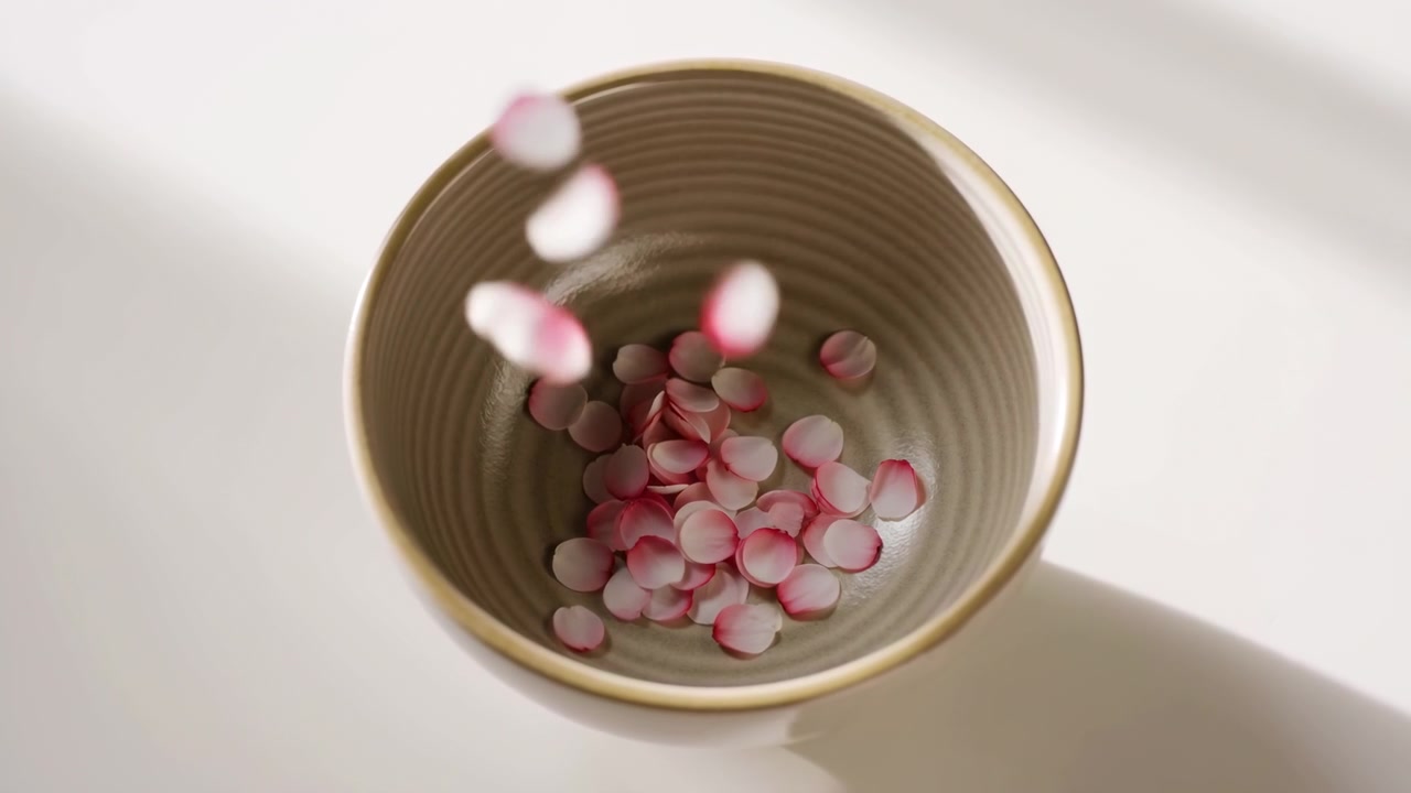 Vista dall'alto di una ciotola di ceramica testurizzata posizionata a sinistra del centro su un tavolo bianco sporco mentre petali di fiori rosa pallido e bianchi cadono e si depositano all'interno