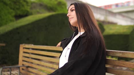 joven mujer bastante caucásica con cabello castaño sentada en un parque en un banco de madera esperando su cita impacientemente seduciendo la expresión de la cara pasando tren verde laberinto nublado configuración lenta