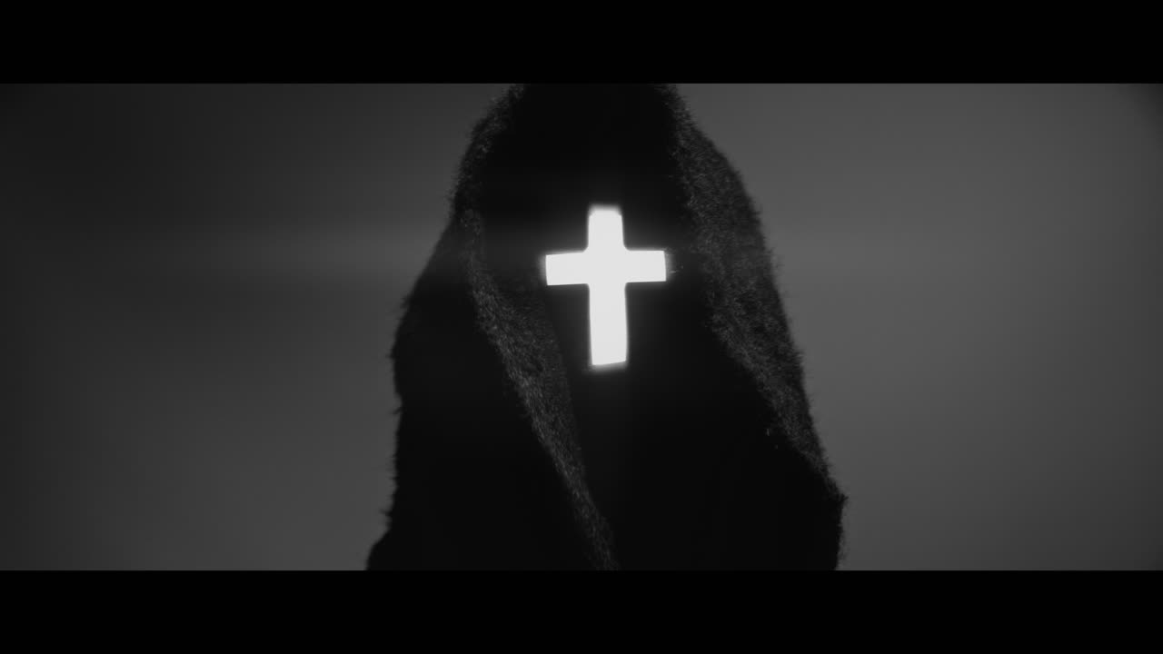 Masked and hooded man with cross of light glowing on his face looks in the camera