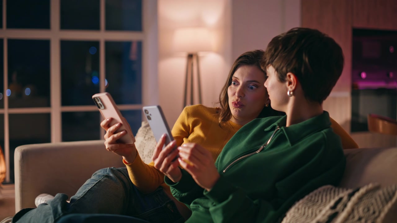 Garotas despreocupadas apreciam o smartphone sentadas em sofá aconchegante de apartamento, close-up