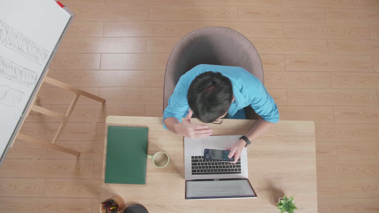 위쪽 뷰  아웃: 화가 난 아시아 남자가 집에 있는 보드 에 있는 나무 책상 에 앉아서 노트북으로 타이핑하는 동안 스마트폰으로 게임을 잃고 있는 장면