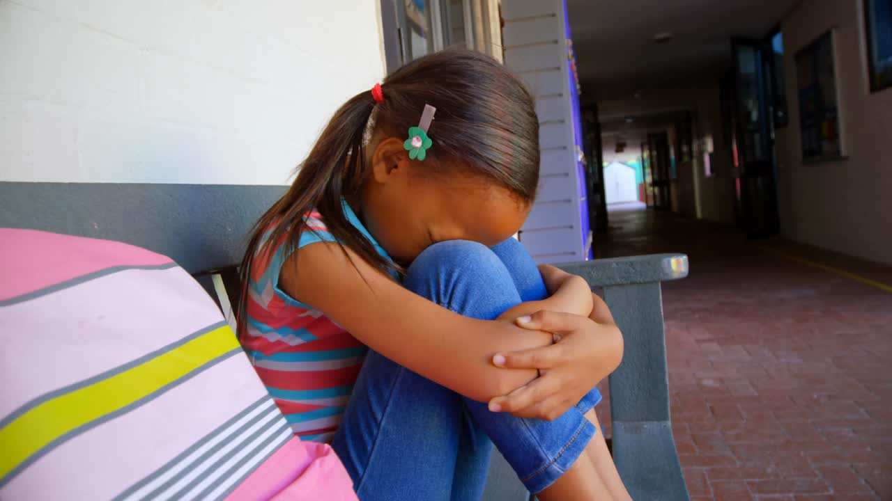 Side view of African American schoolgirl with head down sitting alone on bench in school corridor 4k