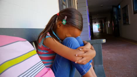 vista lateral de una colegiala afroamericana con la cabeza hacia abajo sentada sola en un banco en el pasillo de la escuela 4k