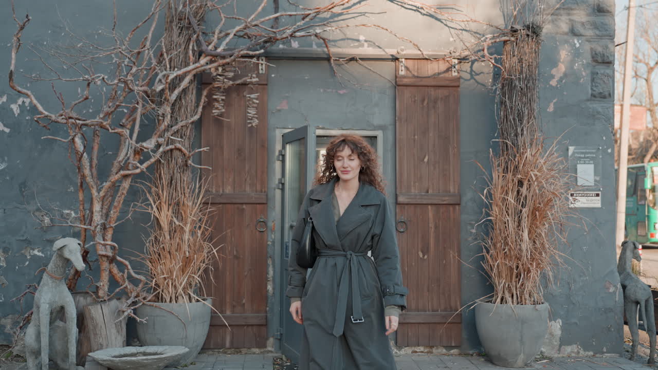 Fiorista caucasica che esce dal negozio, vetrina autunnale, viti, vasi rustici, espressione rilassata, slow living, proprietario di piccola impresa, ritratto in stile catalogo, primo piano esterno, capelli ricci, cappotto, toni neutri.