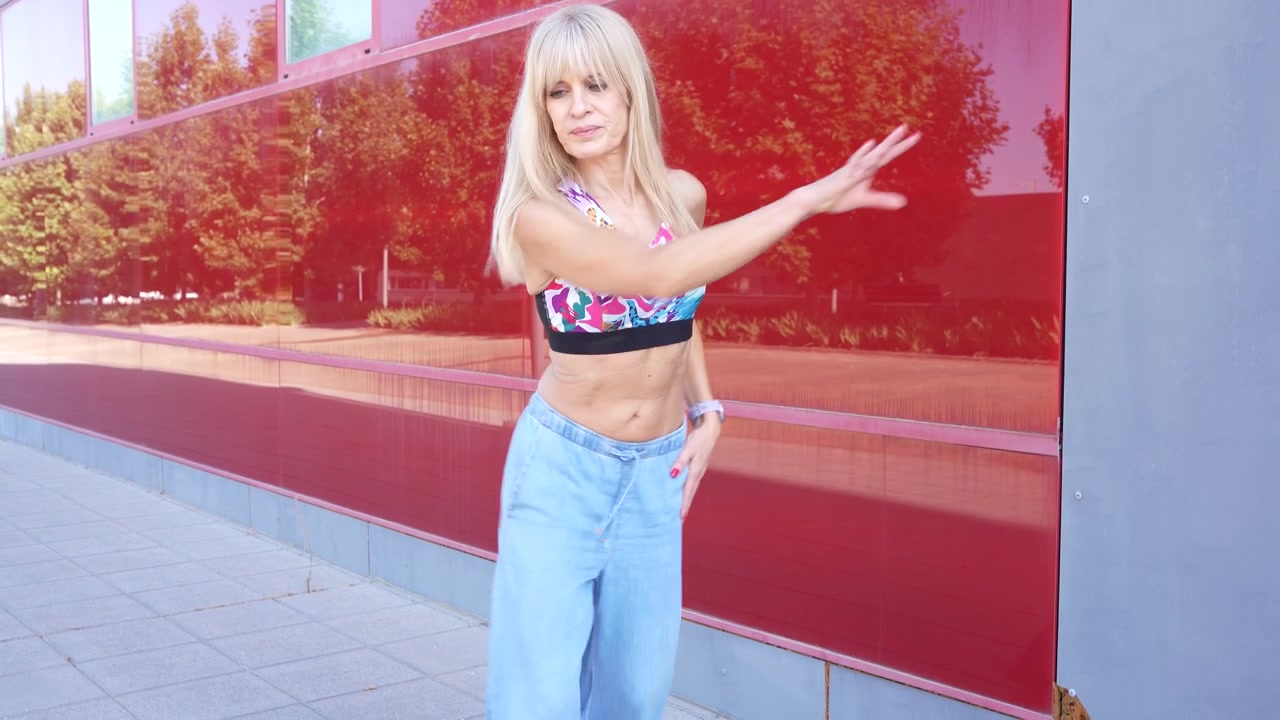 Una giovane donna bionda sta ballando in una vivace strada urbana con un moderno muro rosso di sfondo.