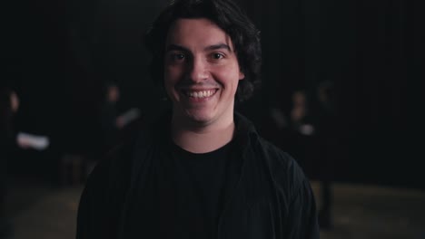 Portrait-of-a-young-confident-brunette-man-in-a-black-suit-the-actor-smiles-and-poses-near-his-colleagues-who-are-preparing-for-a-rehearsal-and-performance-on-stage-in-the-theater