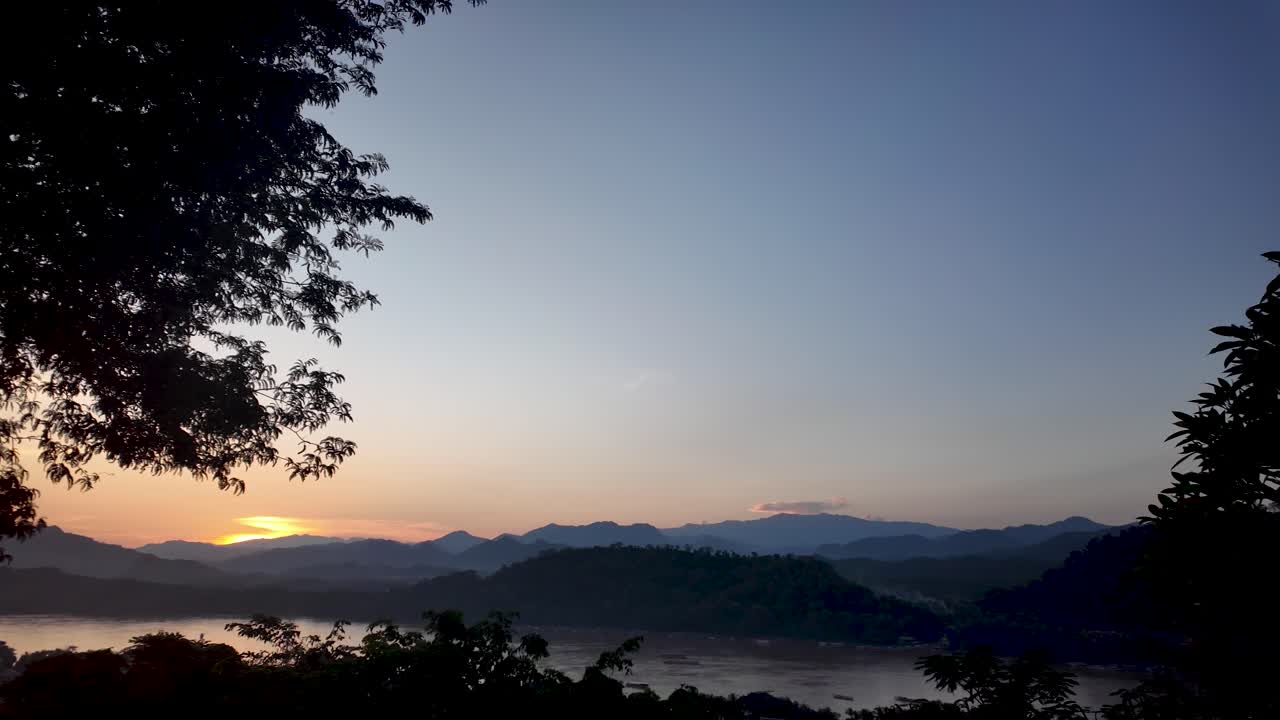 ラオスのプーシー山から見たメコン川に沈むルアンパバーンの夕日の美しい景色。背景には木のシルエットと山並みが広がります。