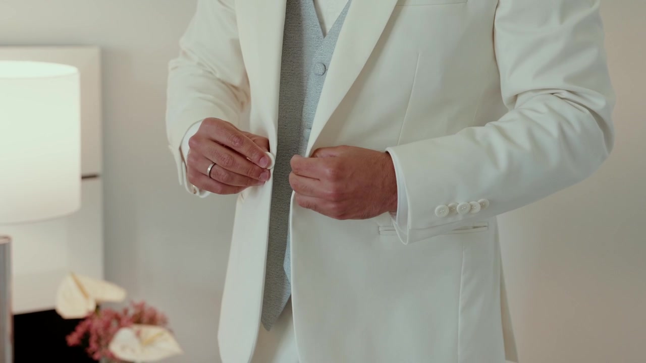 Novio abrochándose un chaleco azul claro debajo de una chaqueta de traje color crema mientras se prepara en el interior