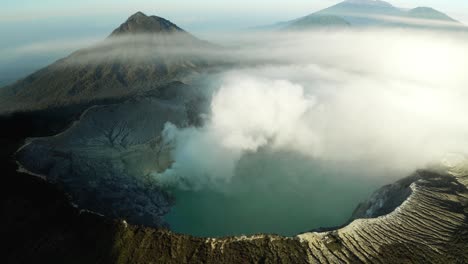 aerial circling the rim of a steaming volcano caldera, with jagged rocks, a turquoise lake, and distant foggy mountain peaks - east java, indonesia